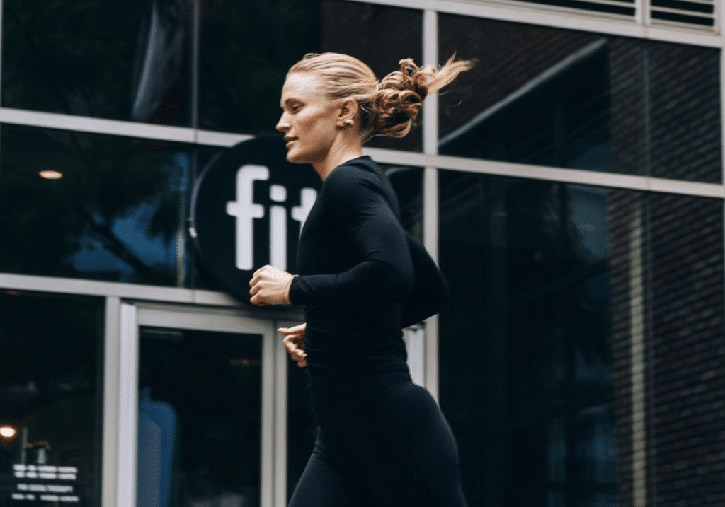 Person in black athletic wear jogging past a building with a "fit" sign in the background.